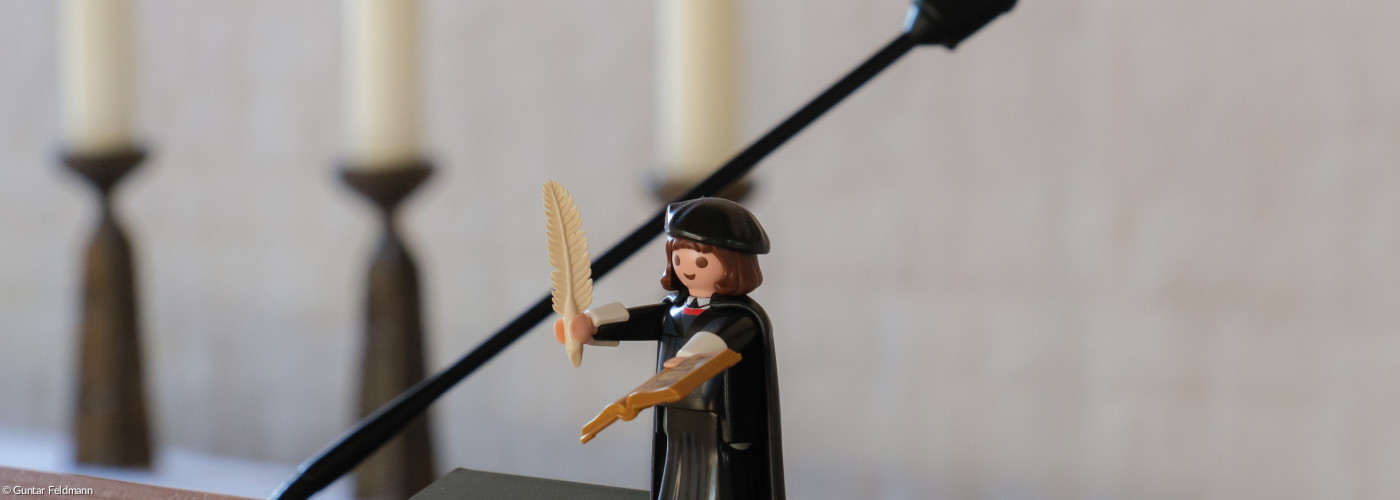 Der Playmobil Luther steht auf dem Ambo im Gottesdienstraum und blickt in Richtung gemeinde.