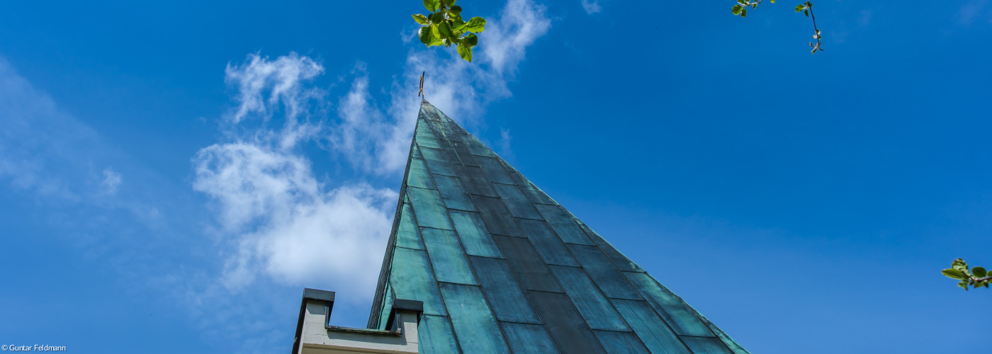 Eine Aufnahme der Thomaskirche steil von unten. Zu sehen ist das spitze Dach und das Kreuz vor dunkelblauem Himmel.