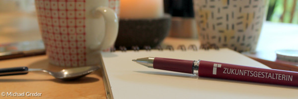 Kaffeetassen, ein Block sowie ein Stift stehen bzw. liegen auf einem Tisch